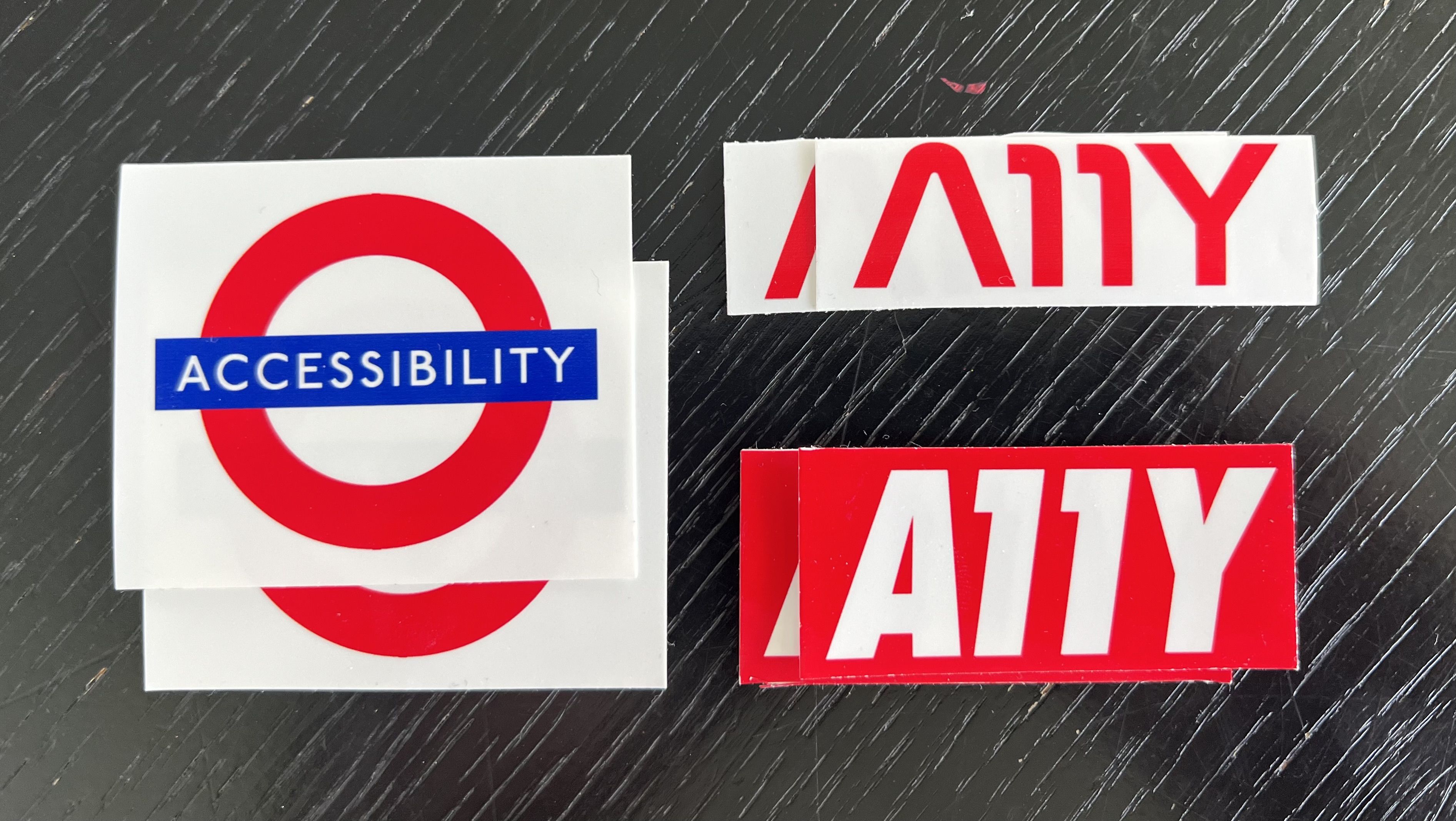 Three self printed and cut stickers. A london underground sign saying Accessibility, A red Nasa typeface text saying A11Y and a OBEY inspired red sticker with white text also saying a11y